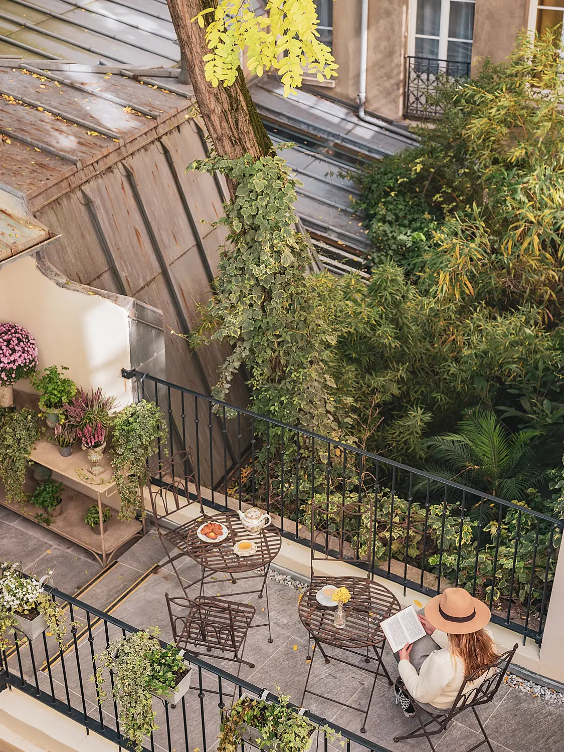 Person reading on a tree-shaded terrace with a wrought-iron balcony.