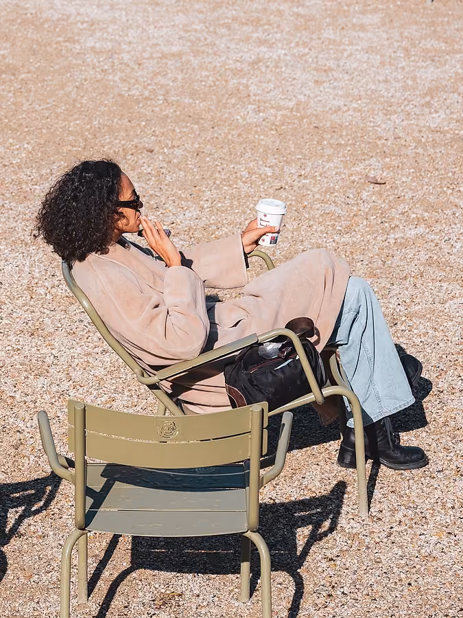 Une femme assise sur une chaise de jardin dans un parc, portant un trench-coat et tenant un gobelet à emporter.