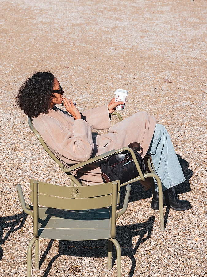 A woman sitting on a garden chair in a park, wearing a trench coat and holding a takeaway cup.