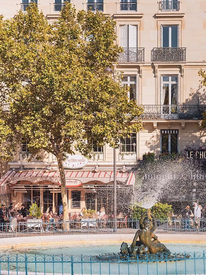 Une vue ensoleillée d'une rue parisienne, centrée sur une fontaine traditionnelle et un vieil immeuble résidentiel.