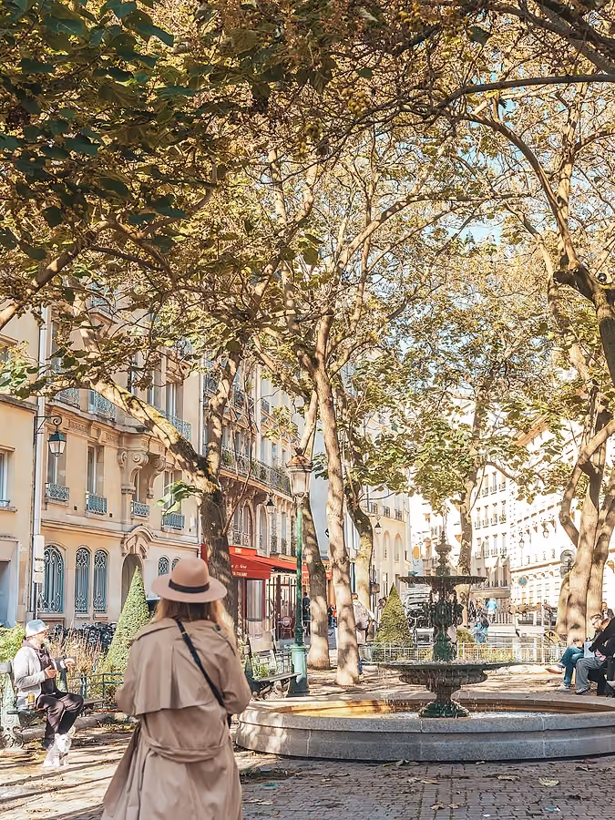 Une femme vue de dos, portant un chapeau et un trench-coat, marchant dans un parc parisien avec une fontaine et des arbres en arrière-plan.