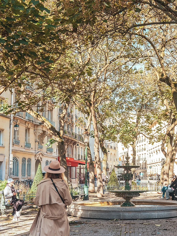 A woman seen from behind, wearing a hat and a trench coat, walking in a Parisian park with a fountain and trees in the background.