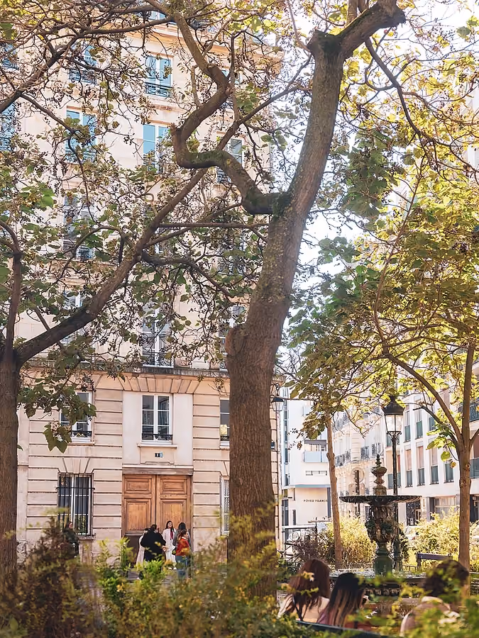 Vue d'une cour parisienne arborée avec une fontaine au premier plan et des personnes marchant près d'un bâtiment classique.