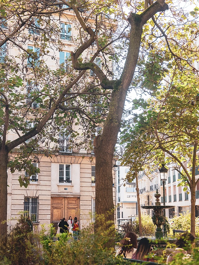 View of a tree-filled Parisian courtyard with a fountain in the foreground and people walking near a classical building.