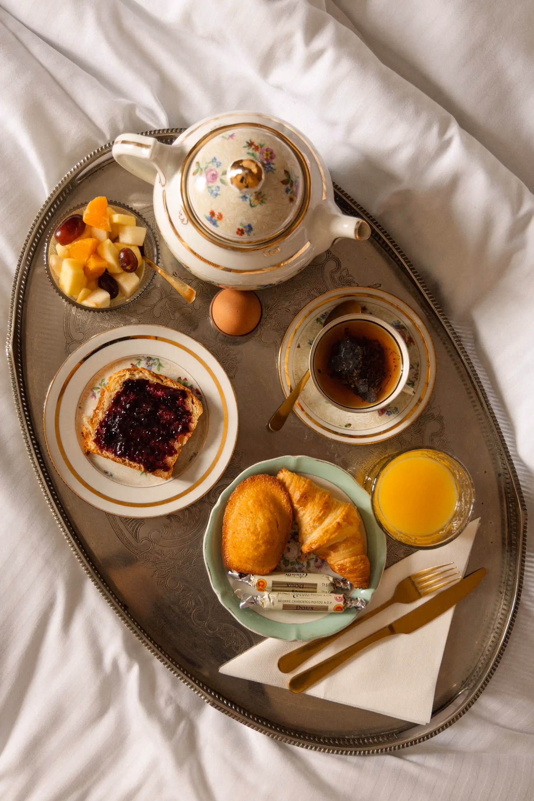 Petit-déjeuner complet servi sur un plateau ovale, composé de thé, viennoiseries et fruits, déposé sur un lit.