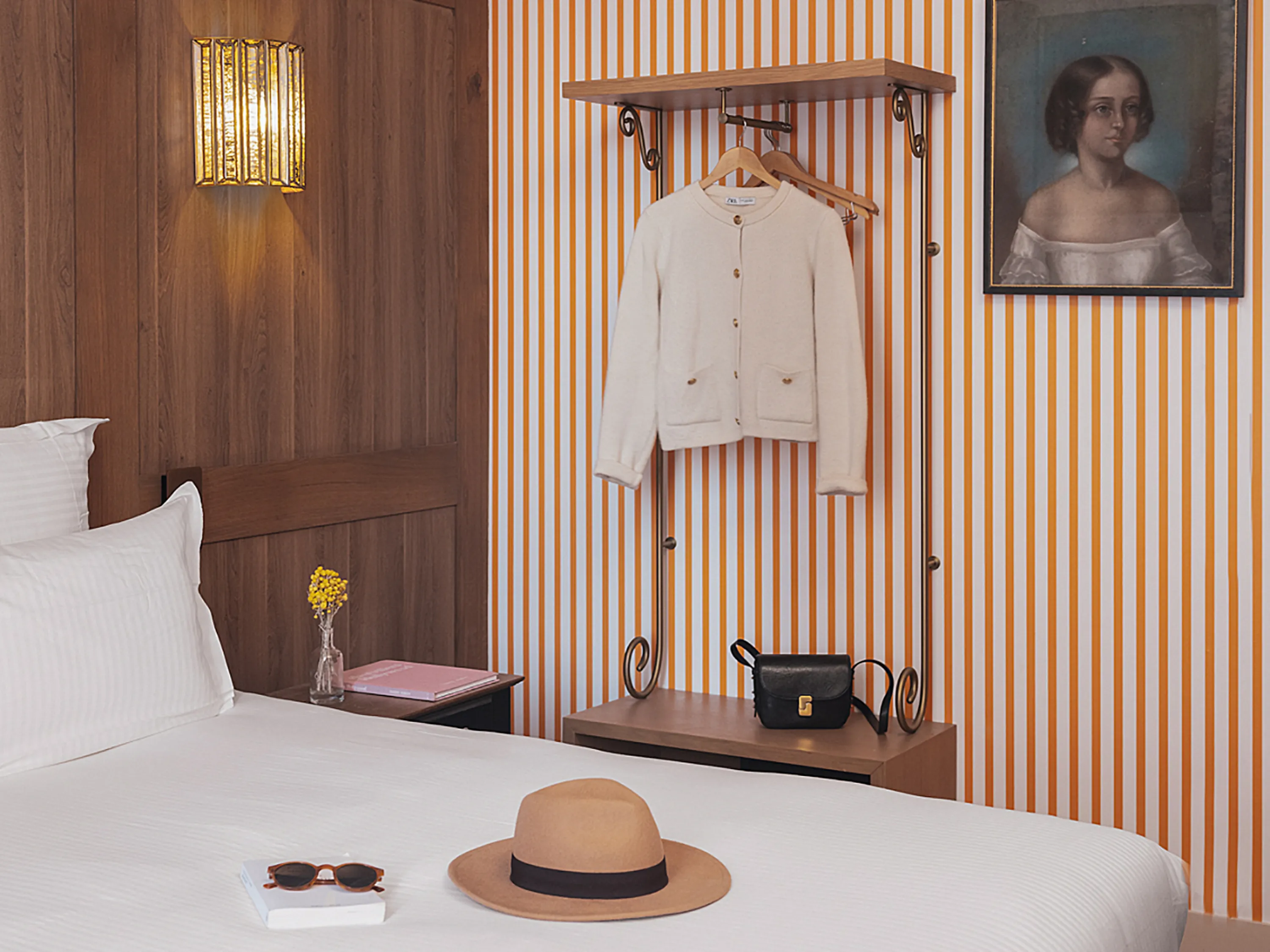 Chambre d'hôtel avec lit, chapeau de paille et portants à vêtements près d'un portrait.