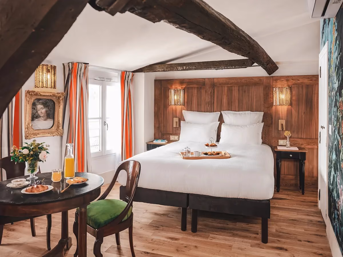 Chambre de l’Hôtel Madeleine de Senlis avec un lit double, des poutres apparentes et une table dressée pour le petit-déjeuner.
