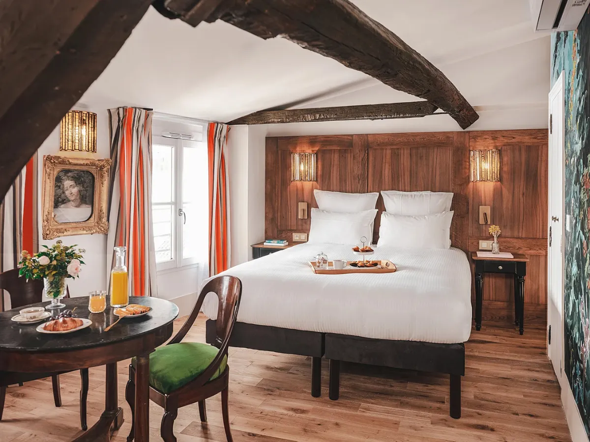 Chambre de l’Hôtel Madeleine de Senlis avec un lit double, des poutres apparentes et une table dressée pour le petit-déjeuner.