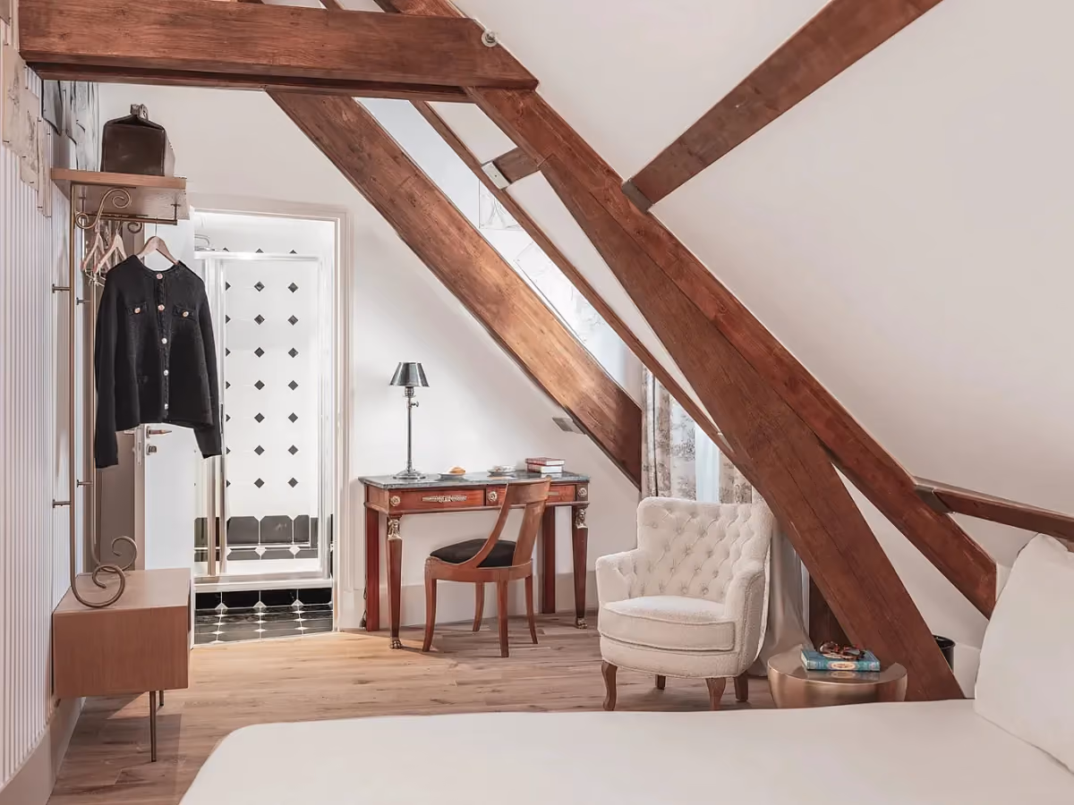 Chambre mansardée avec poutres apparentes, fauteuil et bureau en bois.