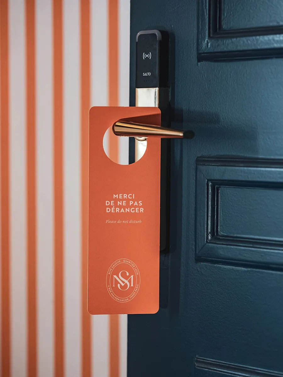 Un panneau de porte « Ne pas déranger » orange avec le logo de l'hôtel NSI, suspendu à une poignée de porte moderne de l’Hôtel Madeleine de Senlis.