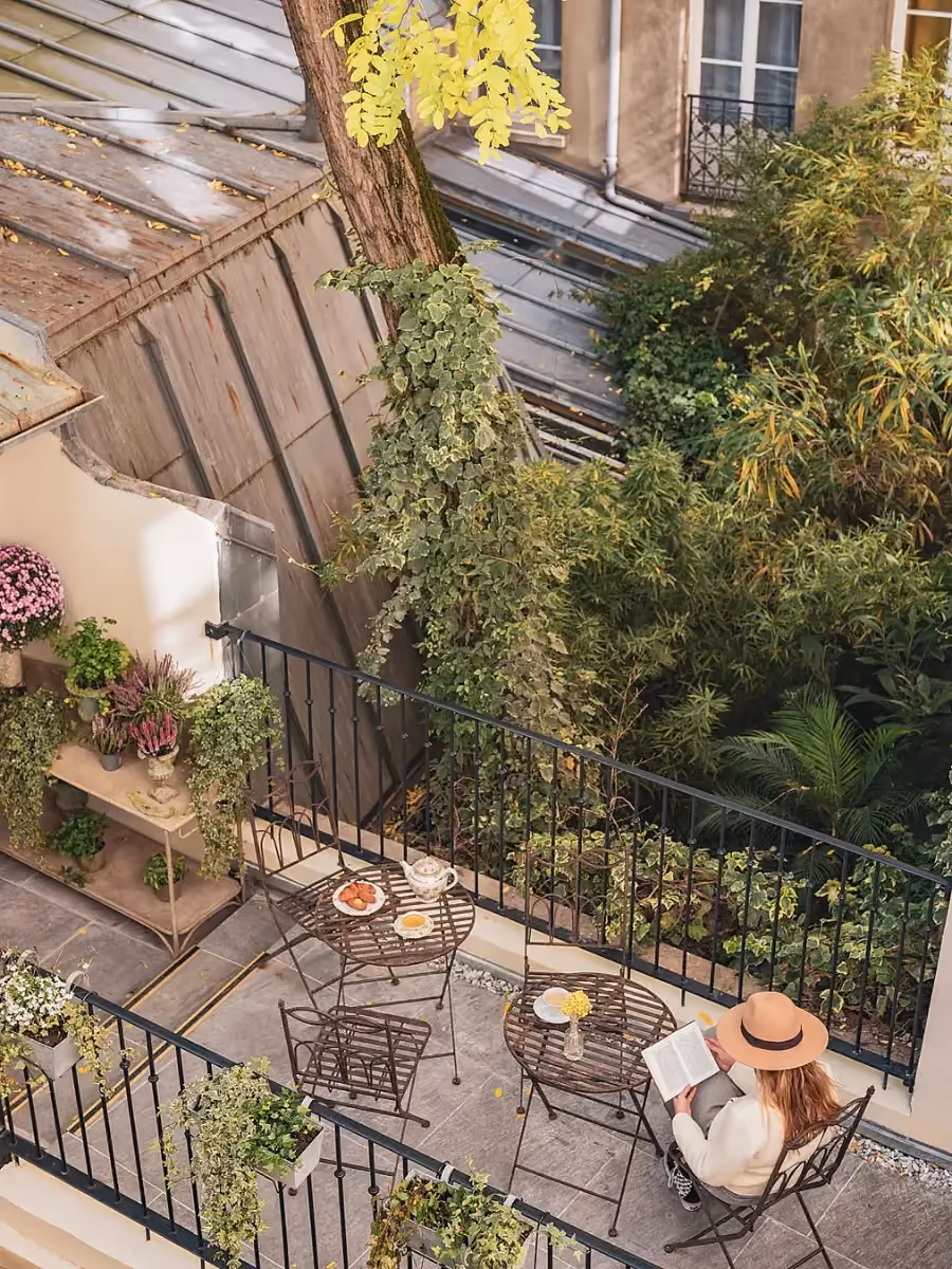 Personne lisant sur la terrasse arborée de l’Hôtel Madeleine de Senlis avec un balcon en fer forgé.