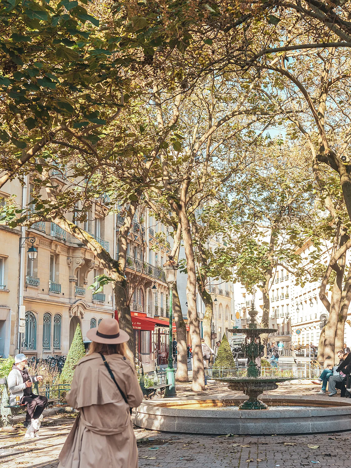 Femme vue de dos portant un chapeau, marchant dans un parc parisien avec une fontaine ancienne et des arbres.