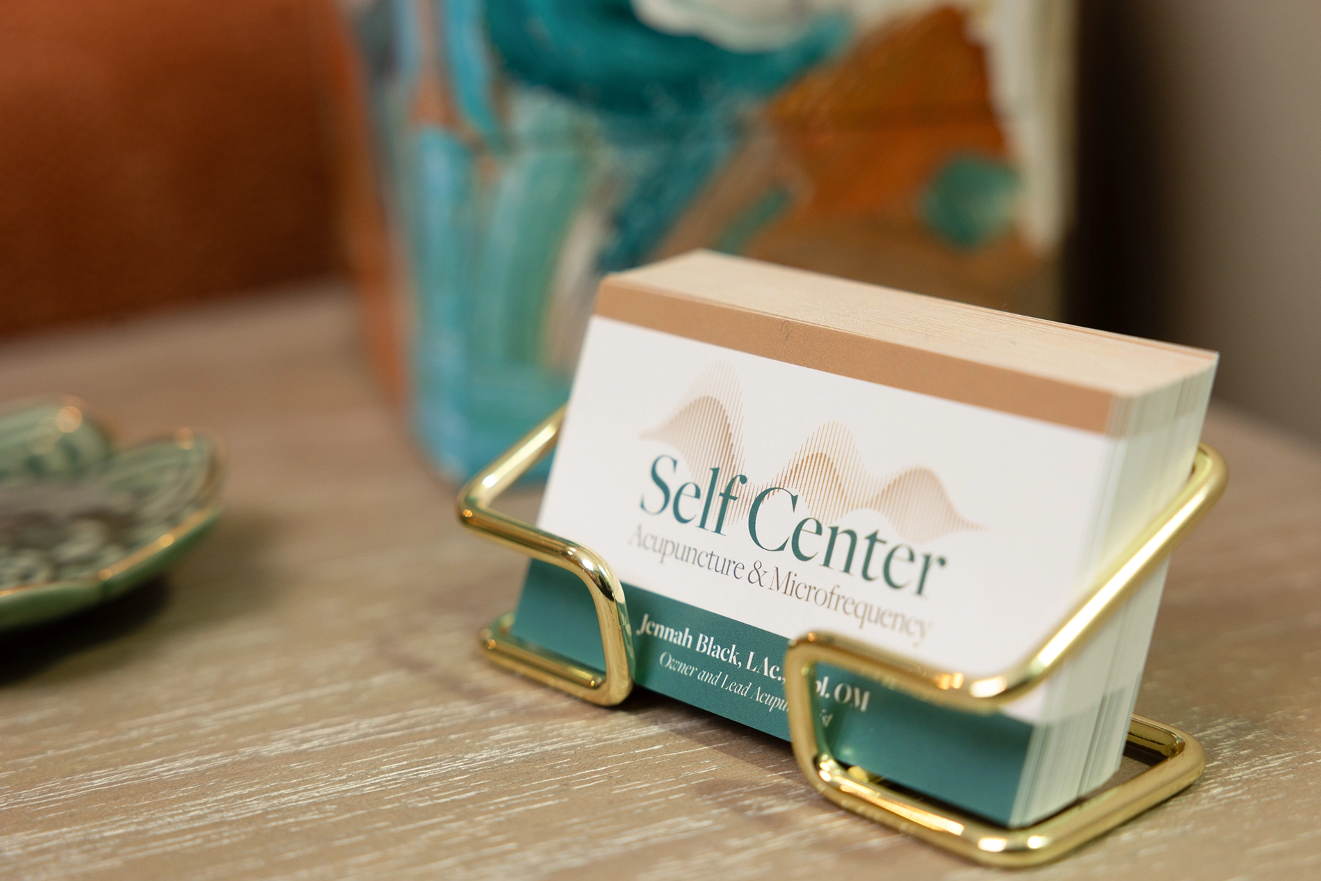 Stack of Self Center acupuncture and microfrequency business cards held in a gold metal holder on a wooden surface with blurred decorative items in the background.