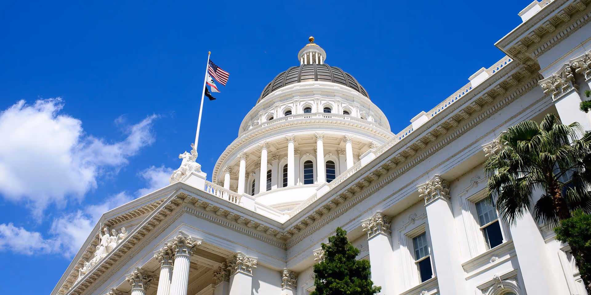 The California State Capitol dome with American and California flags flying against a blue sky.