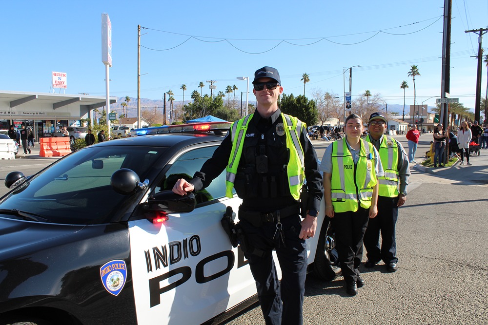 Three police officers wearing reflective vests standing beside an Indio police car on a sunny street.