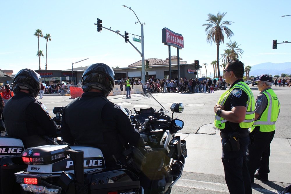 Police officers on motorcycles and on foot monitoring a crowd at a street intersection on a sunny day.