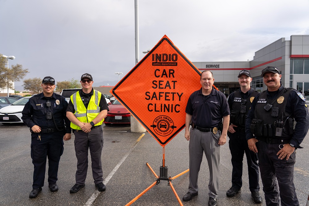 Five police officers standing beside an orange sign reading Car Seat Safety Clinic in a parking lot.