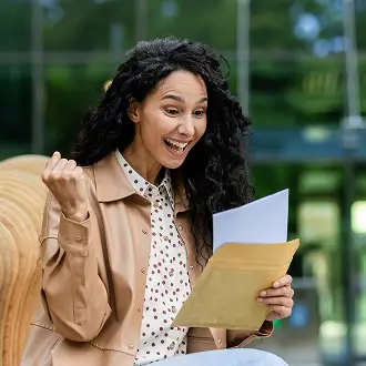 Excited woman with curly hair holding an envelope and letter outside a modern building.