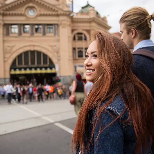 Smiling woman with long reddish hair and denim jacket standing near a man in front of a historic building with a crowd in the background.
