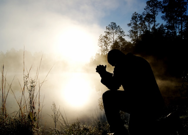A silhouette of a person kneeling in prayer against a backdrop of a bright, misty sunrise or sunset with tall trees in the distance.