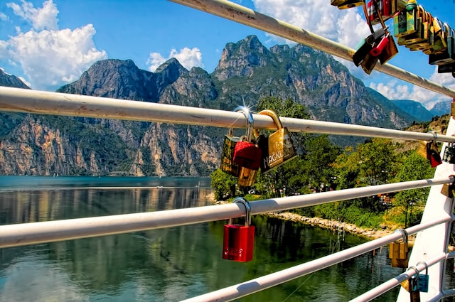 A photograph of numerous padlocks, including a prominent red one, attached to the metal railing of a walkway overlooking a large lake and steep, rocky mountains under a blue sky.