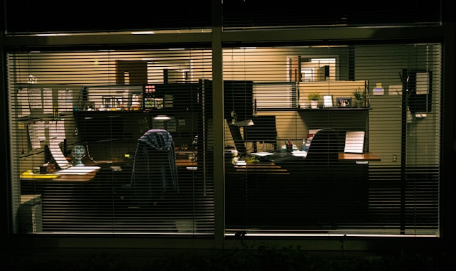 A photograph taken through a window into a dark, empty office at night, showing unoccupied desks, computer monitors, and chairs behind horizontal blinds.