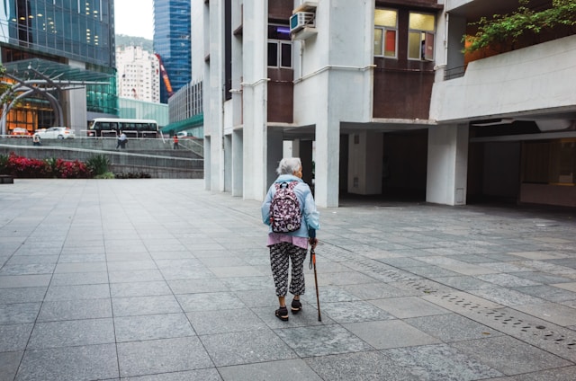 A photograph of an elderly person seen from behind, walking slowly with a cane across a wide, empty stone plaza toward modern city buildings in the distance.