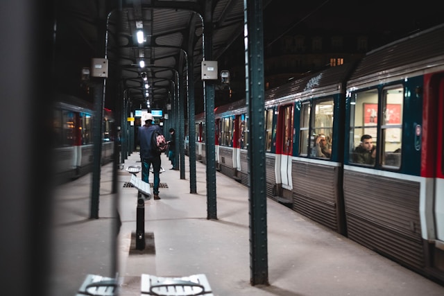 A blurred foreground view looking down a train platform at night, with a person walking toward a stationary train and light reflecting off the platform floor.