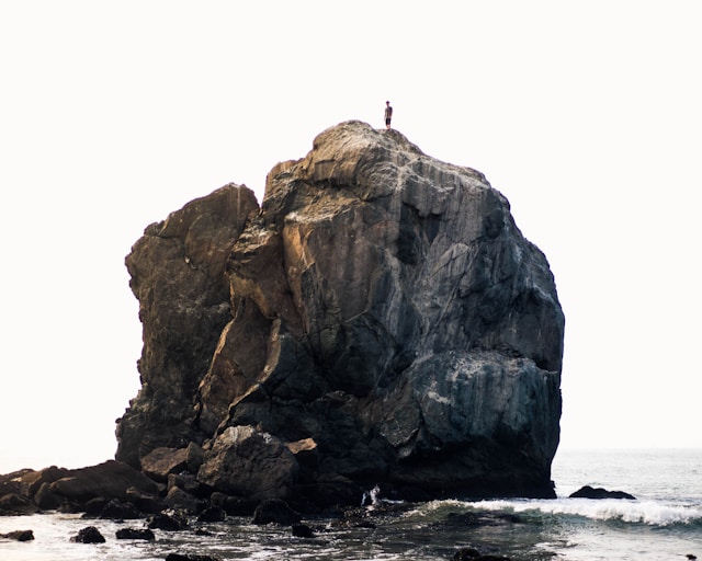 A large, jagged rock formation rises out of the ocean under a pale, overcast sky. A tiny silhouette of a person stands on the highest peak of the rock.
