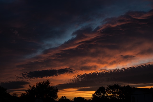 A dramatic twilight sky filled with heavy, textured clouds. The clouds are illuminated from below in vibrant shades of orange and yellow, transitioning into deep purple and dark blue hues higher up. The dark silhouettes of trees and a building line the bottom edge against the glowing horizon.