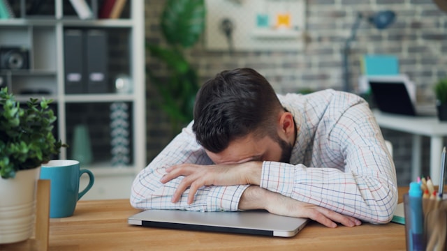 A tired man in a checkered shirt resting his head on his folded arms over a closed laptop at an office desk.