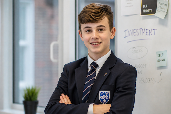 Smiling teenage boy in a school uniform with crossed arms standing in front of a whiteboard with notes about investment and success.
