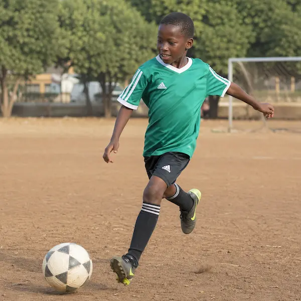 Young boy in green jersey kicking a soccer ball on a dirt field with a goal and trees in the background.