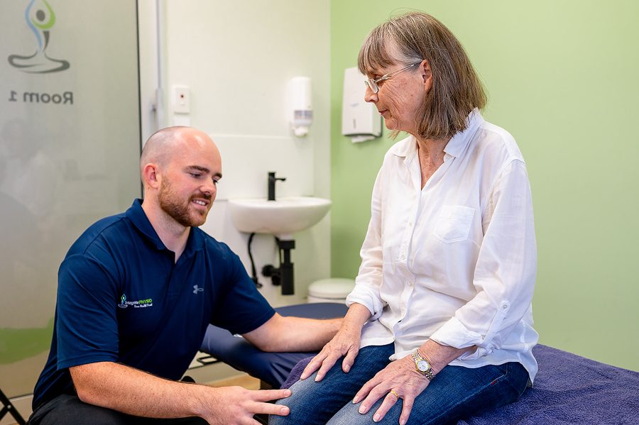Physiotherapist assessing an elderly woman's knee in a clinical treatment room.