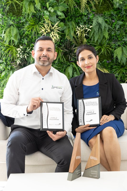 Saurabh Bhojwani and Yamini Kapoor seated holding framed Business & Innovation awards with two wooden trophies on a table in front of them.