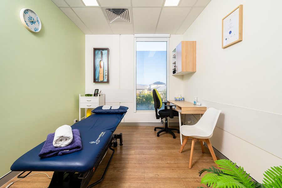 Integrate Physio bright treatment room with blue massage table, white chair, wooden floor, and desk by a window.
