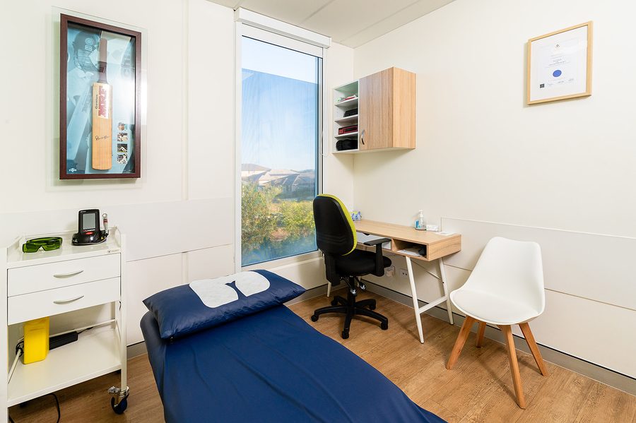 Integrate Physio bright medical consultation room with examination bed, office chair, white chair, framed cricket bat, and large window.