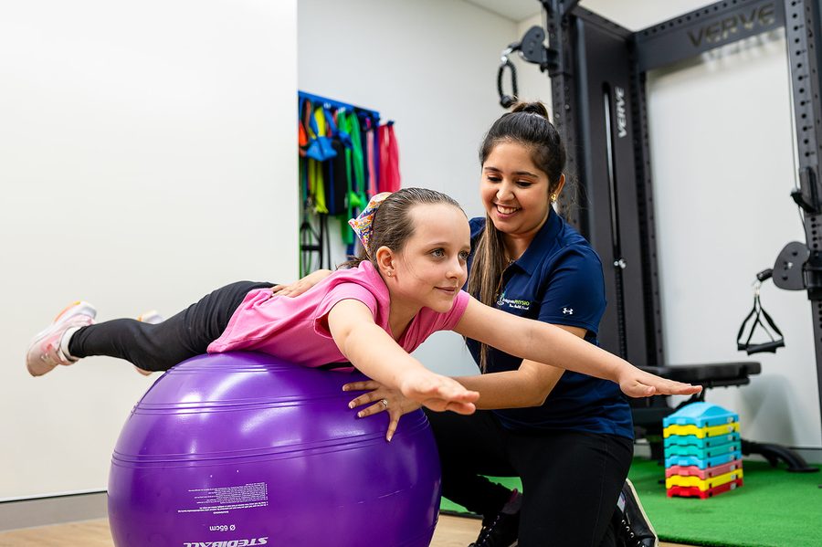 Child in pink shirt balancing face down on a purple exercise ball while guided by a smiling trainer in a gym.