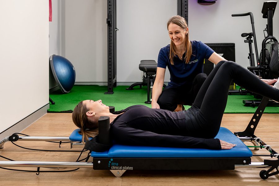 Woman exercises on Pilates reformer machine guided by a smiling instructor in a gym studio.