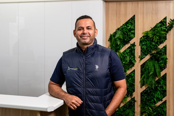 Saurabh Bhojwani in a navy vest and shirt leaning on a white countertop with a background of green plants on wood panels.