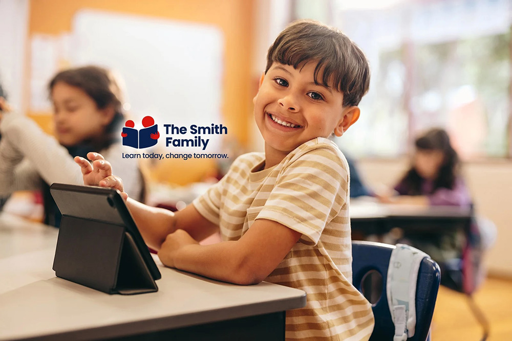Smiling child sitting at desk using a tablet in a classroom with other students in the background.