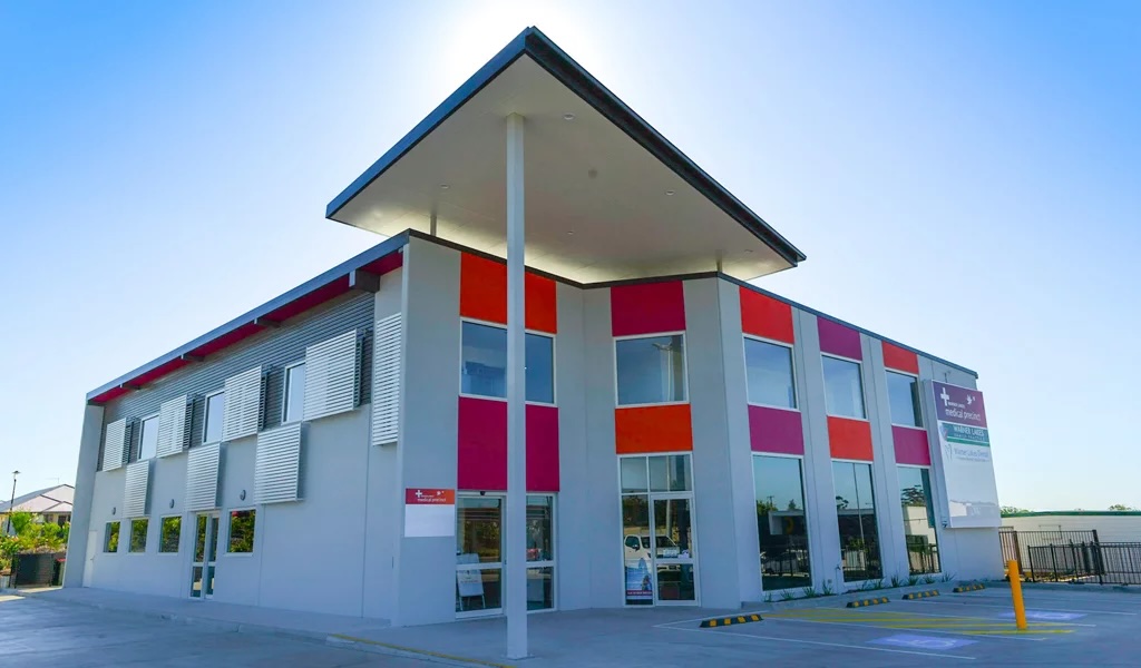 Modern two-storey medical centre building with grey walls, red and orange panels, and large windows under a clear blue sky.