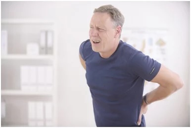 Man in a blue shirt grimacing while holding his lower back in pain indoors.