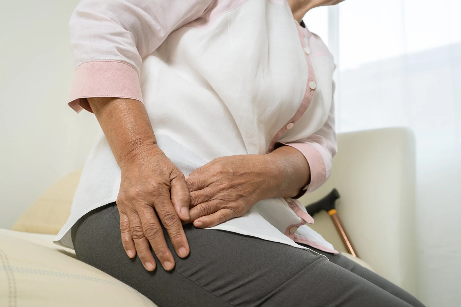 Older person sitting and holding their hip in pain with a walking cane in the background.