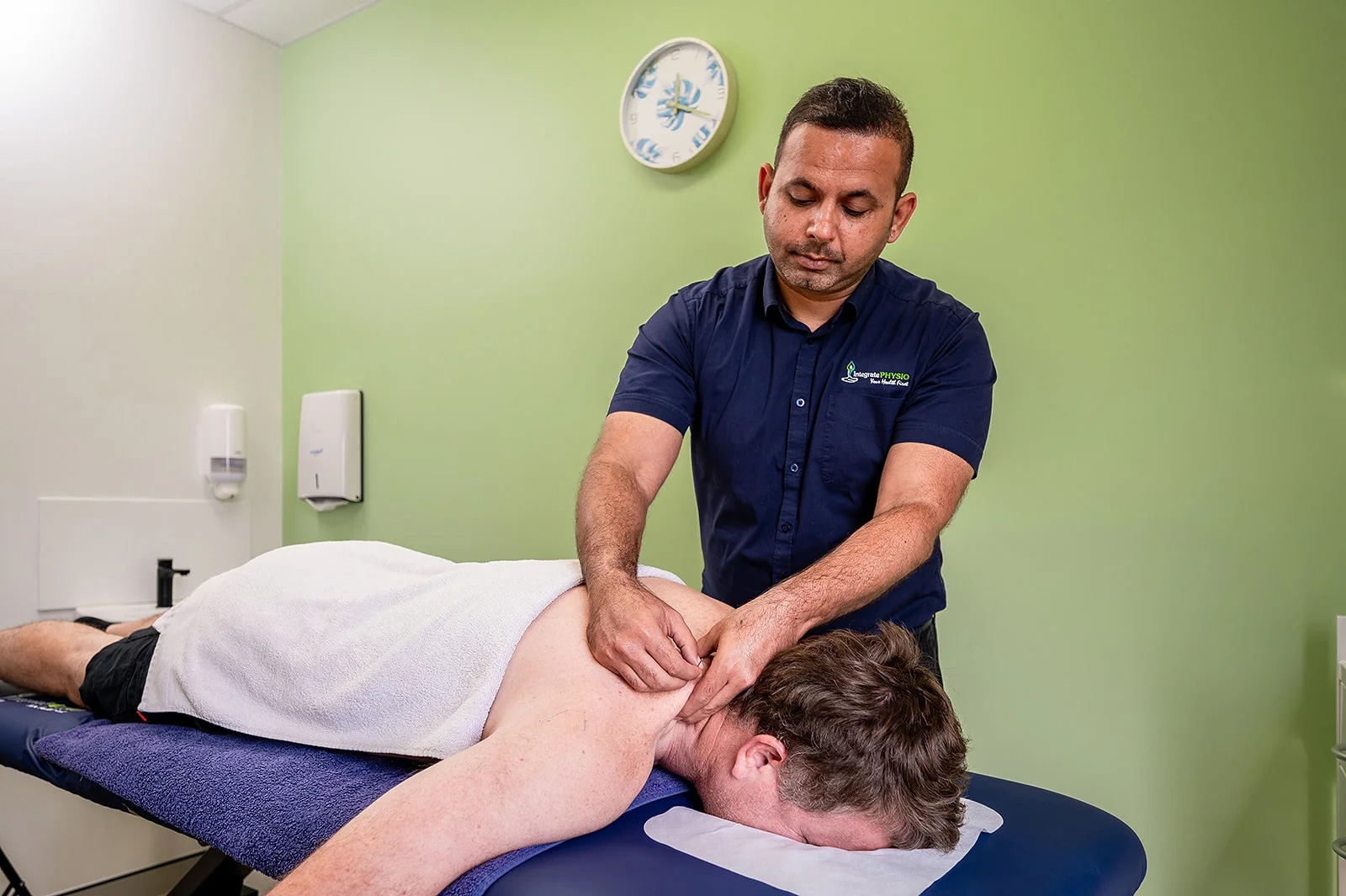 Sam Bhojwani performing physiotheray on a man's upper back in a clinic room.