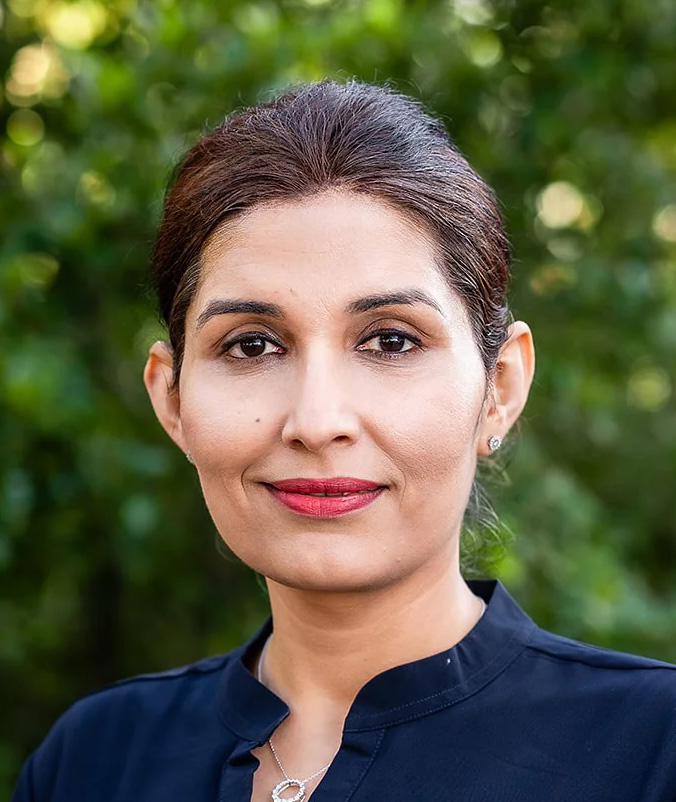 Portrait of a Yamini Kapoor with dark hair tied back, wearing a navy top and silver necklace, with a green blurred background.