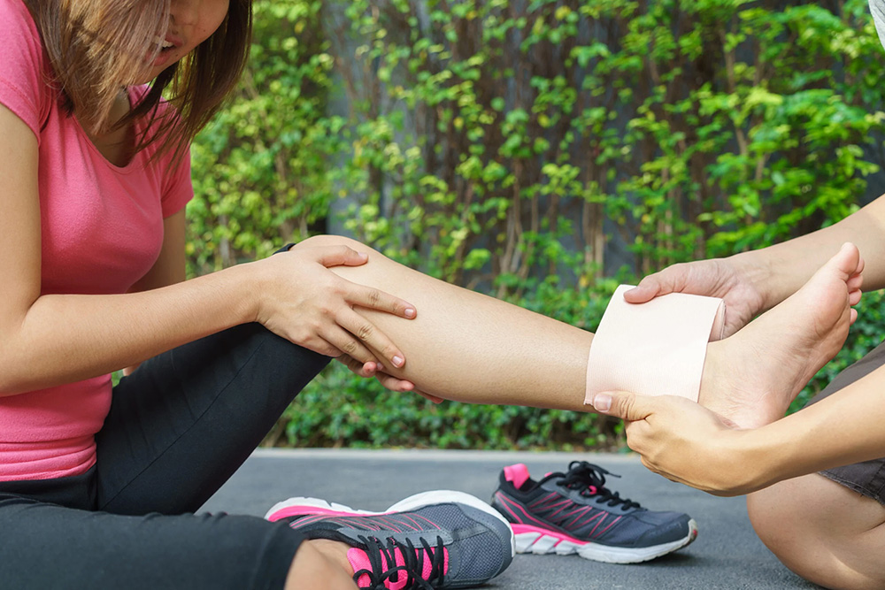 Person in pink shirt holding their injured leg while another person wraps an ankle support band.