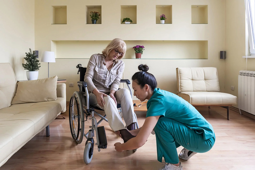 Caregiver in teal scrubs kneeling to assist elderly woman in wheelchair with putting on a sock.