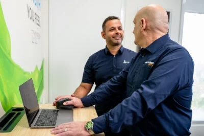 Two men in navy blue shirts smiling and discussing while using a laptop on a standing desk.