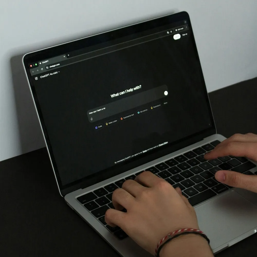 Hände, die auf einem Laptop mit geöffneter ChatGPT-Webseite tippen, Anzeige eines dunklen Chatfensters mit dem Satz 'What can I help with?'